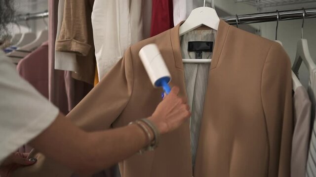 Garment care of contemporary clothes at home. Spbd Black woman cleans beige coat from dirt with lint roller in well-equipped closet in light room closeup