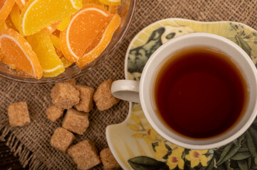Multicolored shaped marmalade lemon slices in a glass vase, pieces of brown cane sugar and a cup of tea on a saucer, close-up, selective focus.