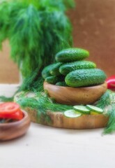 On a wooden background, there is a large bunch of green dill in a jar, and a pile of green cucumbers in a plate