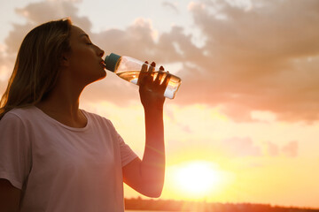 Young woman drinking water to prevent heat stroke outdoors at sunset, space for text