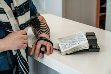 Religious adult jew in a tallit and tefillin on his left hand stands on a table with an open...