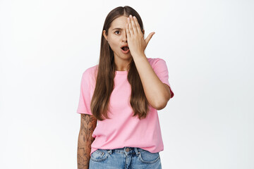 Woman with hand on half of face, looks with one eye with shocked, worried expression, standing against white background