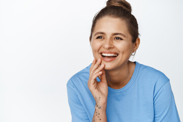 Close up of candid modern girl laughing, smiling and touching her face while looking aside, standing in blue tshirt against white background