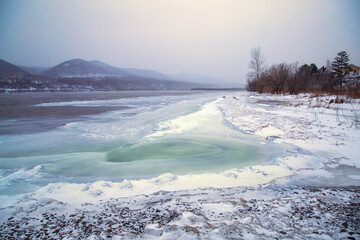 Winter landscape with an ice-covered river, snow-capped banks and mountains in the background.