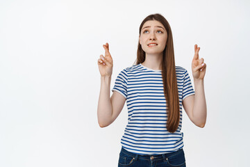 Hopeful girl prays, cross fingers and looking up nervously, making wish against white background