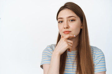 Fototapeta premium Thoughtful girl looking at camera with hand on chin, thinking, listening to smth interesting, white background