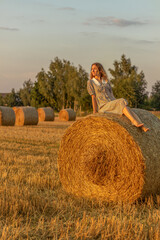 Naklejka na ściany i meble Young woman in blue dress close up on the field background with haystacks at sunset time.