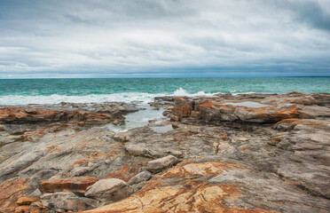 Big boulders of red rock and sea waves against the dramatic sky background