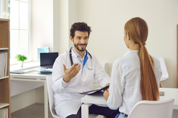 Obraz premium Smiling positive male doctor sitting at desk in his office and talking to female patient. Young woman or teen girl listening to her physician during consultation in modern clinic or hospital
