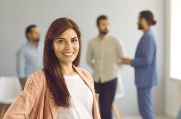 Close up portrait of a successful young business woman looking at camera with confident expression. Smiling woman on a background of people talking to each other in a bright room. Confidence concept.