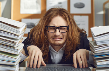 Unhappy tired employee doing paperwork and working on laptop computer. Portrait of funny sad...