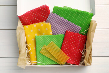 Colorful beeswax food wraps in box on white wooden table, top view