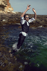 man with a beard and sunglasses in clothes a vest and a white shirt jump in sea among the rocks in summer