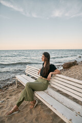 Beautiful brunette woman looking far into the sea horizon. Girls sits on white bench on the beach. Concept of work-life balance and wellbeing.