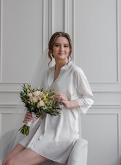 A beautiful young girl in a dressing gown at the window with a bouquet in her hands is a blonde with hair and makeup in a white room.