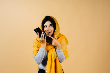 Obraz premium Smiling young arabian muslim woman in hijab light clothes posing isolated on yellow background. People religious Islam lifestyle concept. copy space. Hold mobile phone sending audio message