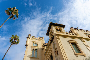 Perspective view with a view from below of a large and luxurious house in Arabic style, with large windows, and with palm trees in the background with the blue cloudy sky.