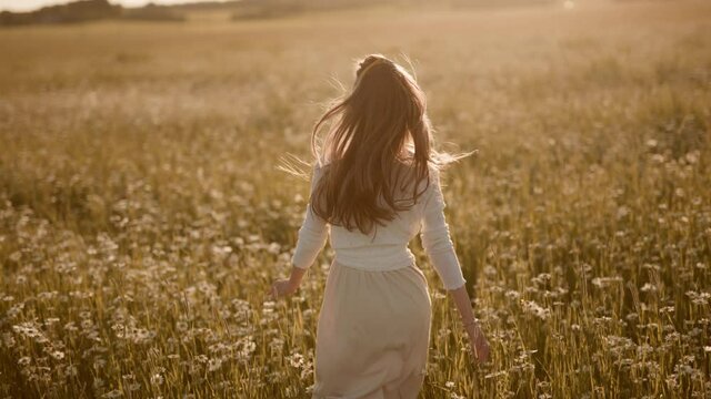 A happy woman is running in slow motion across field with camomiles at sunset. Carefree girl is enjoying freedom and calmness in nature during vacations. Concept of recreation and happiness or summer.