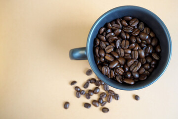 on the right, a dark mug filled with roasted whole coffee beans stands on a beige background of a table with scattered coffee beans. top view