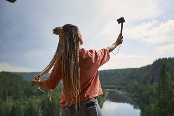 a modern girl with dreadlocks records her blog standing with her back to the camera on top of a cliff overlooking the river and dense forest. High quality photo
