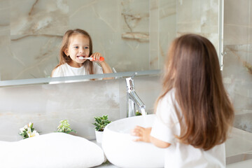 A happy little girl brushes her teeth in front of a mirror in the bathroom. Morning routine. Hygiene.