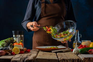 The chef prepares a Greek salad and places it from a large glass bowl into a serving plate. Lots of ingredients. Book of recipes. Step by step recipe. Vitamins. - Powered by Adobe