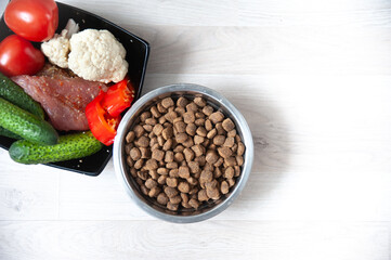 Bowl with dog food, next to natural products, vegetables and meat close-up and space for text, top view. Healthy pet food concept