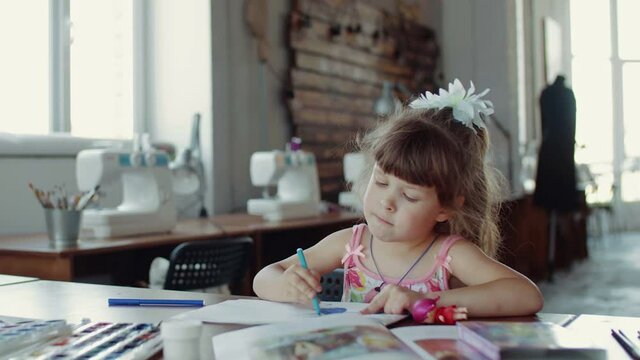 A little girl is sitting at a table and drawing on paper with different colors and brushes