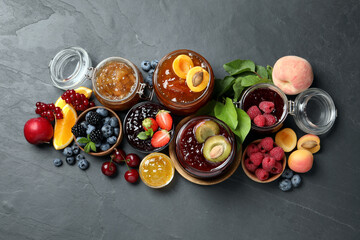 Jars with different jams and fresh fruits on black table, flat lay