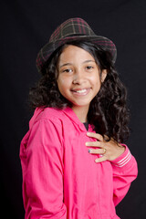 
Curly hair latin girl wearing a hat smiles with hand on chest, black background
