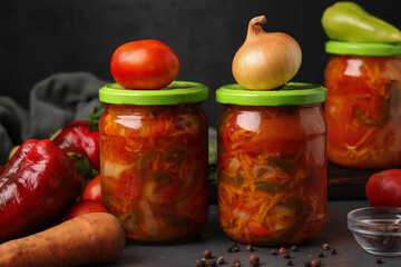 Vegetable salad of tomatoes, carrots, onions and peppers in jars for the winter on dark gray background