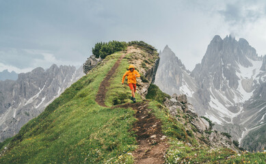 Dressed bright orange softshell jacket runner running by green mountain path with picturesque Dolomite Alps range background,. Active people and European mountain hiking tourism concept image.