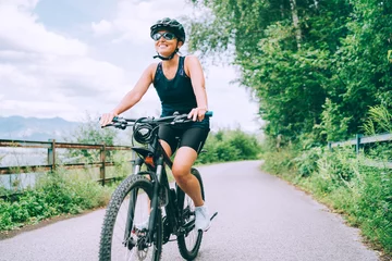 Tuinposter Portrait of a happy smiling woman dressed in cycling clothes, helmet and sunglasses riding a bicycle on the asphalt out-of-town bicycle path. Active sporty people concept image. © Soloviova Liudmyla