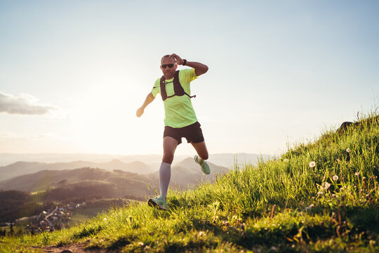 Active mountain trail runner dressed bright t-shirt with a backpack in sports sunglasses running endurance ultramarathon race by picturesque hills at sunset time. Sporty active people concept image..