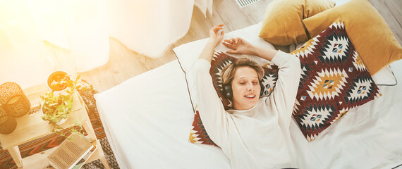 Smiling young female lying on cozy bed dressed pajamas listening to music using wireless headphones. Audio playing modern technology and free time spending concept