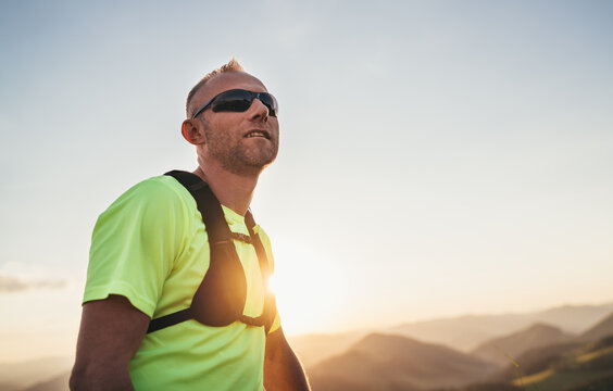 Portrait of Active mountain trail runner dressed bright t-shirt with backpack and sport sunglasses resting while he sky running by picturesque hills at sunset time. Sporty active people concept image