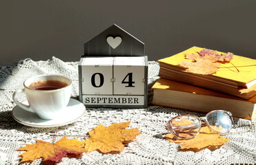 Calendar for September 4 : the name of the month in English, cubes with the numbers 0 and 4, a cup of tea, books, maple leaves on a gray openwork napkin, side view, gray background