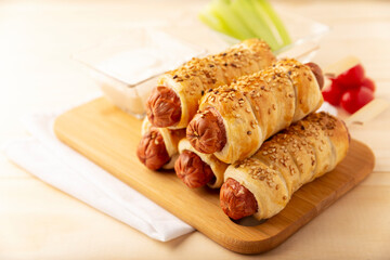 delicious snack of sausages wrapped in puff pastry with sesame seeds accompanied by cherry tomatoes, celery and sauce. Served on wooden board