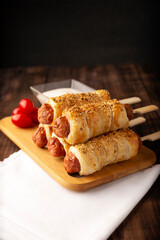 delicious snack of sausages wrapped in puff pastry with sesame seeds accompanied by cherry tomatoes and sauce. Served on wooden board