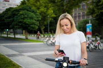 Fototapeta na wymiar Smiling woman renting electric kick scooter at city street. Modern sharing urban transport concept.
