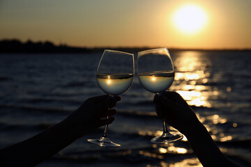 Women clinking glasses of wine near river at sunset, closeup