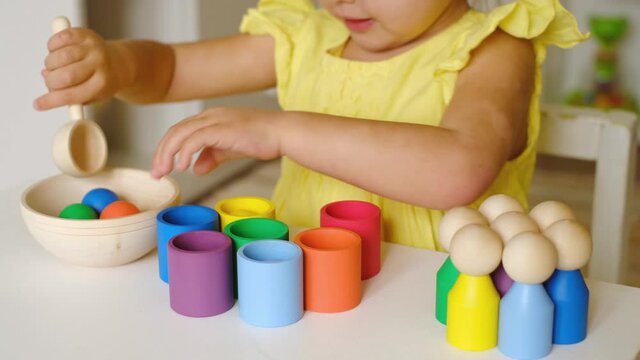 A two-year-old child puts colored balls in a spoon and throws them into cups of the corresponding color. The child learns colors. A sorter toy for studying flowers and preschool children