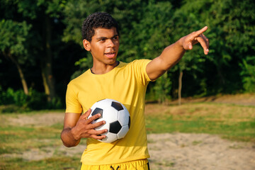 Young smiling Brazilian man holds a ball and pointing away with his finger before playing on a grassy sandy beach in summer. 