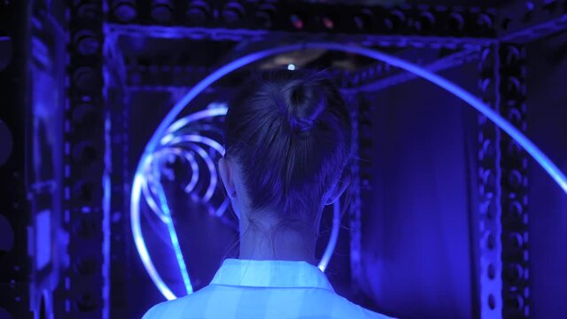 Back view of young woman looking around at interactive futuristic exhibition or museum with blue illumination. Immersive, sci-fi and technology concept