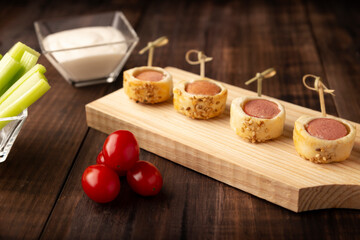 delicious snack of sliced sausages wrapped and in puff pastry with sesame seeds accompanied by cherry tomatoes, celery sticks and sauce. Served on wooden board