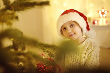 Little boy wearing Santa hat ready for celebrate Christmas. Cute child decorating the Christmas tree with glass toy. Baby hopes of magic and gifts at New Year night.