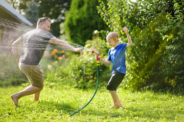 Funny little boy with his father playing with garden hose in sunny backyard. Preschooler child...