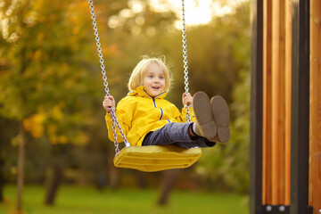 Little boy having fun on a swing on the playground in public park on autumn day. Happy child enjoy swinging. Active outdoors leisure for child - obrazy, fototapety, plakaty