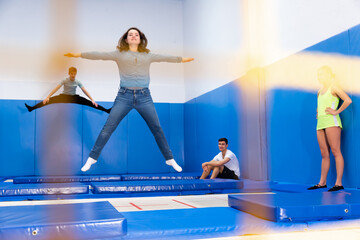Concentrated young people jumping on trampolines behind safety net, fulfilling jump movements during training..