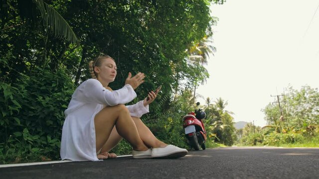Woman on red motorbike in white clothes drive on forest road trail trip. One girl caucasian tourist sit, relax, rest, near scooter. Use a smartphone, take a selfie. Motorcycle rent.